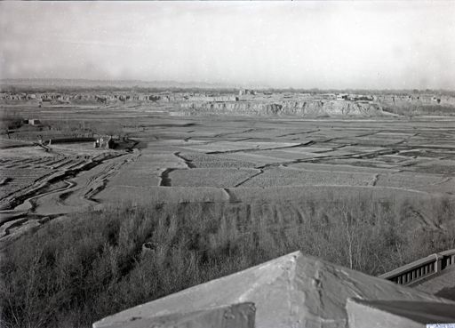 B 1235: Blick von einem Dachgiebel: mit Feldern, kleinem christlichen Friedhof und einem Plateau mit Häusern bis zum Gebirge.