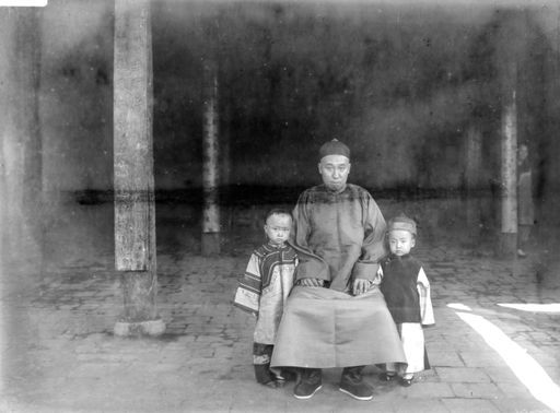 B 1581: Ein Mann in chinesischer Tracht sitzt neben seinem kleinen Sohn und seiner kleinen Tochter in einer großen Halle mit Pfeilern, die Chinesische Schrift tragen. Im Hintergrund rechts verbirgt sich eine Frau, vielleicht die Mutter.