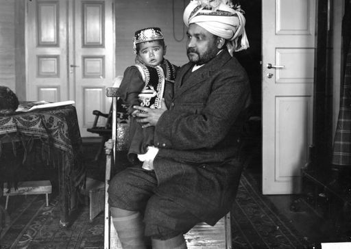 B 1667: Mann mit Turban und in europäischer Kleidung in Zimmer mit westlicher Einrichtung. In seinem Arm trägt er kleinen Jungen in eleganter Volkstracht.- vermutlich der kashmirische Aksakal in Maralbashi.