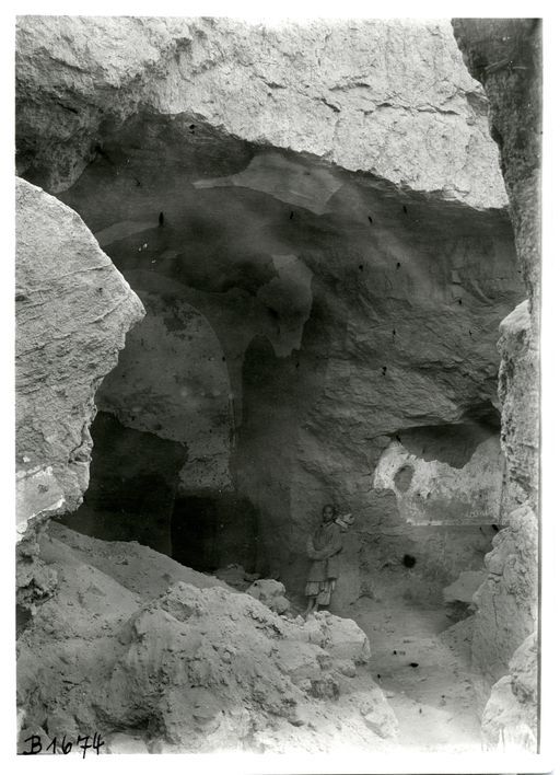 B 1674: Cella einer Höhle mit Tonnengewölbe und Umwandlungsgang, geweißte Verputzreste. Blick durch zerstörte Front von links. Rechts zeigt sich ein ähnlicher Nebenraum wie links. Junge lehnt an der rechten Wand.