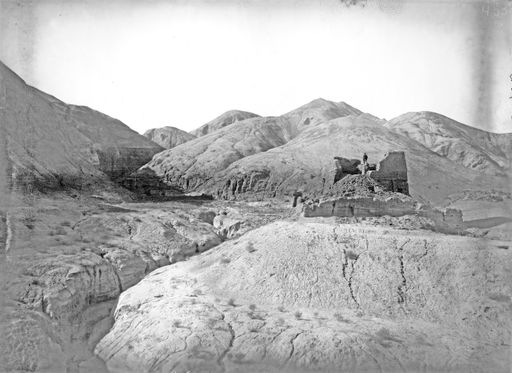 B1702: Th. Bartus steht auf Stūpa-Stumpf in zerklüftetem Tal vor Hügeln mit Höhleneingängen, darüber vielleicht die Ruinem von B 1692 (B_1702_Murtuk_II_Anlage.jpg)