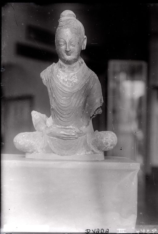 B 1909: Sitzender Buddha, sehr beschädigt. Im Hintergrund Ausstellung im Museum für Völkerkunde