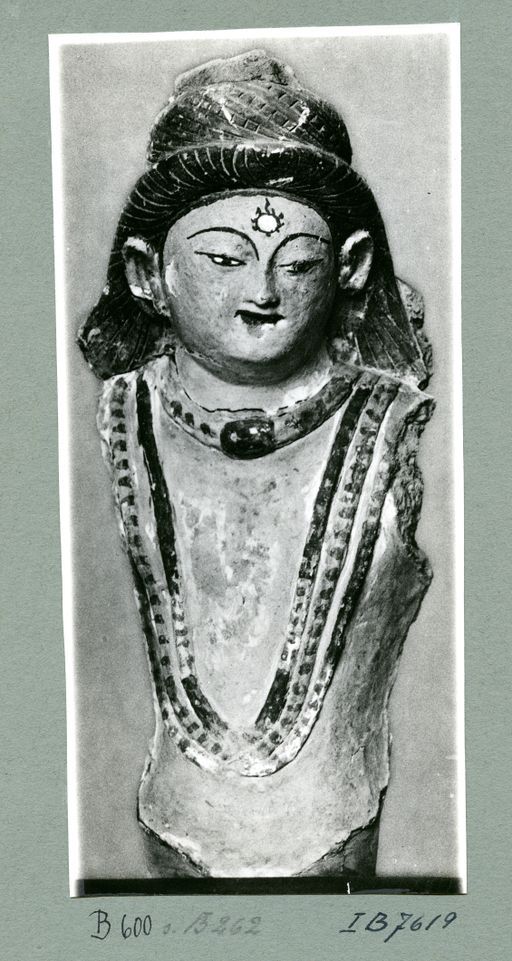 B 600: Büste eines Bodhisattva mit flammender Ūrṇā auf der Stirn. Er hat schwarzes langes Haar und ein rotes Perlendiadem, sowie drei rot, grün, ocker bemalte Ketten und einen Halsring mit Amulettbüchse; Reste von Gold