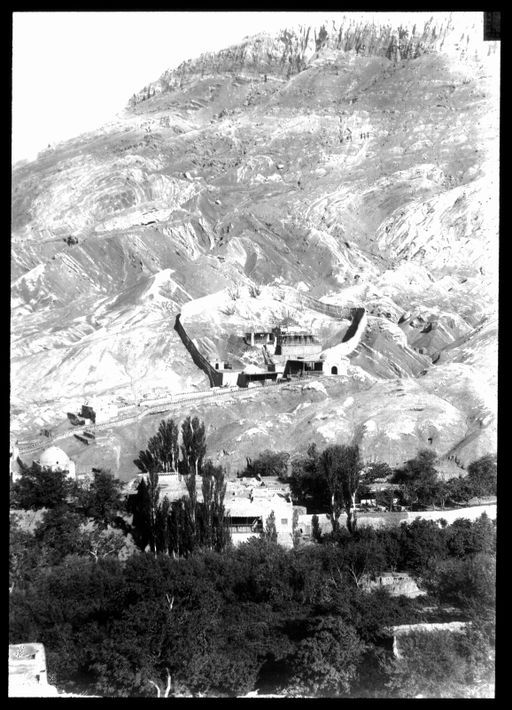 T 17: Moschee der Siebenschläfer: Toyok Mazar. Blick aus dem modernen Dorf auf die ummauerte Anlage am Bergabhang und buddhistische Höhlen am Feldrain