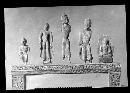 T 410: Fünf kleine hölzerne Buddhastatuetten (3 stehend, einer europäisch sitzend, einer im Meditationssitz) auf einem gemusterten Rahmen